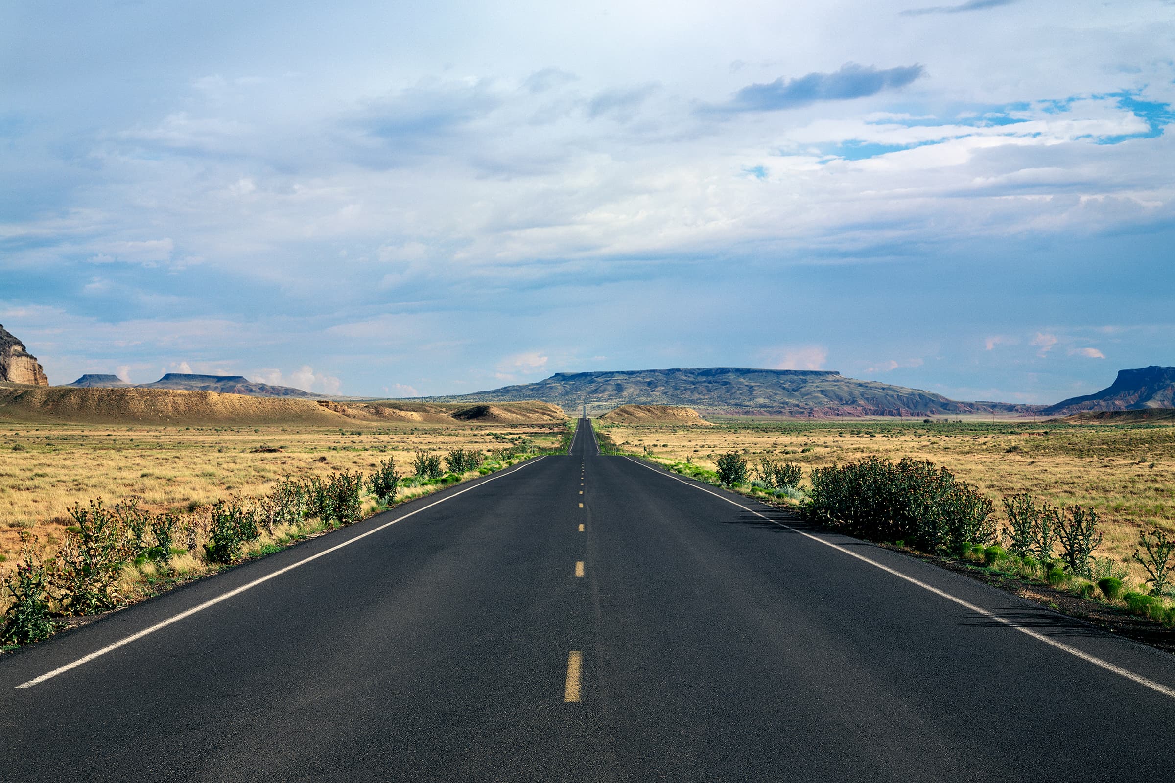 A color photograph of an empty two-lane highway running straight from the foreground to the horizon through flat, golden desert grassland, toward a long, flat-topped mesa under a wide, cloudy blue sky.