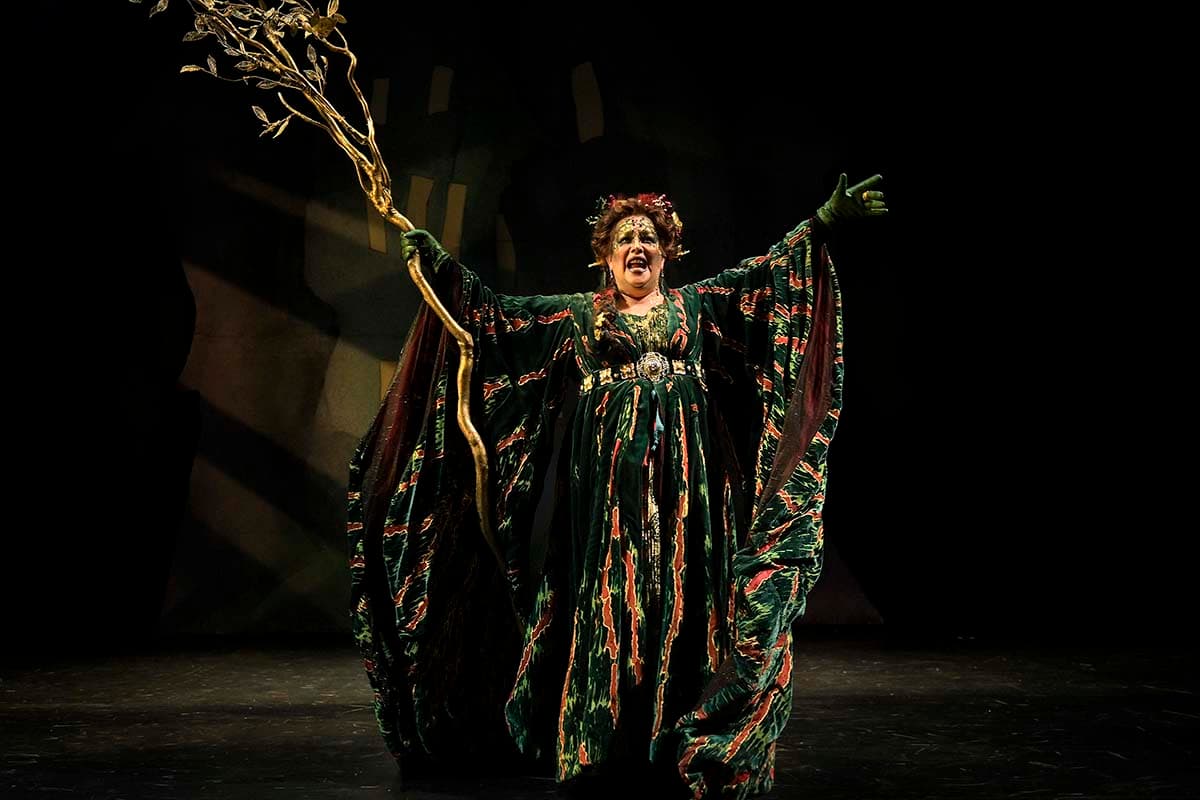 A performer stands center stage under a spotlight against a dark background. She wears an elaborate, flowing green gown featuring red and orange tree-bark or moss-like textures and wide, draped sleeves. She has her arms outstretched wide, holding a tall, golden tree-branch staff in her right hand while delivering a dramatic expression.