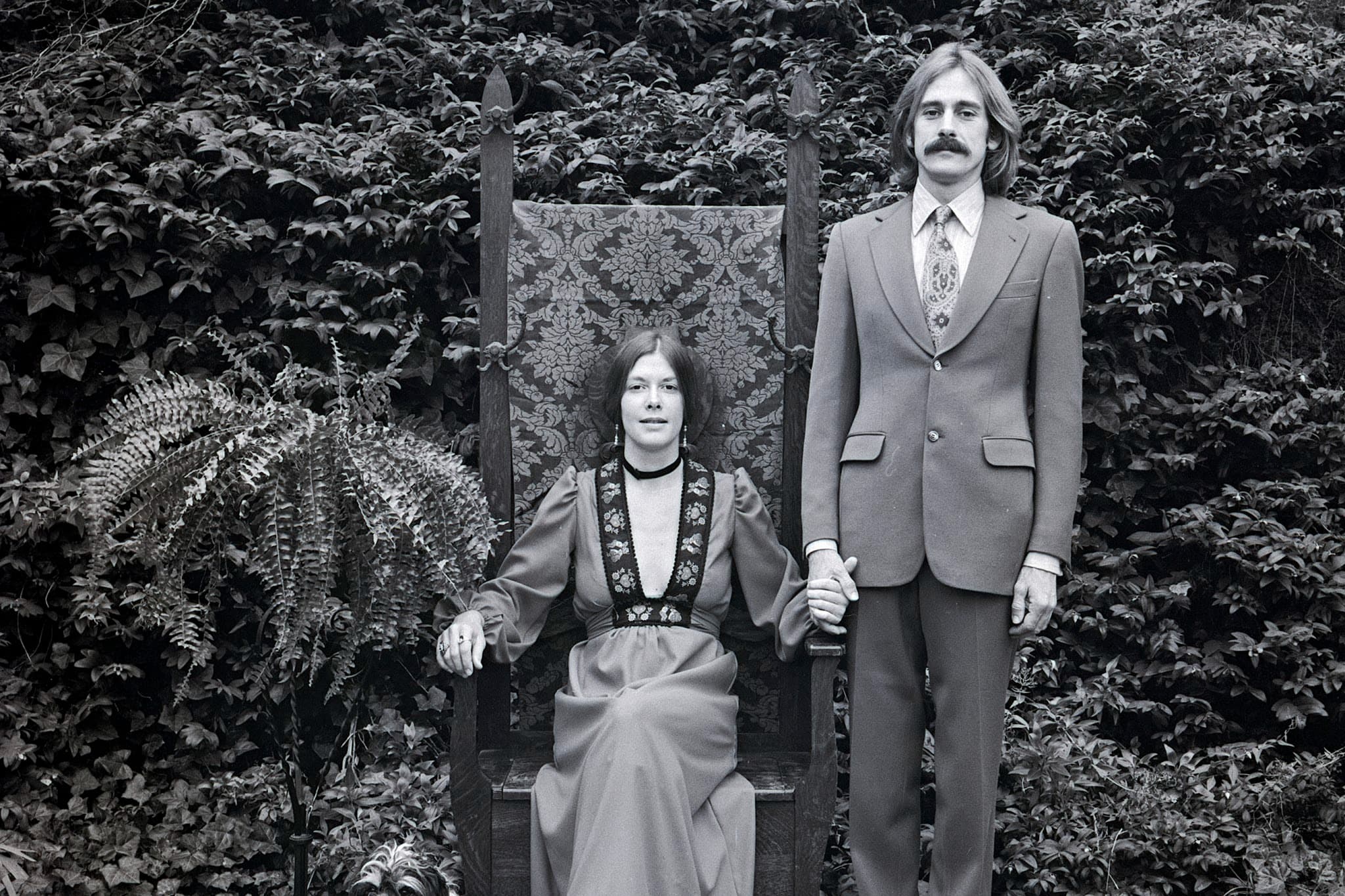 Couple with fern outdoors. A woman with long hair parted down the middle sits in a tall, high-backed wooden chair covered in a floral damask fabric. She wears a long dress with an embroidered V-neckline and a black choker. Standing next to her, a man with a mustache and shoulder-length hair wears a suit jacket, patterned tie, and trousers. They are holding hands, posed against a dense backdrop of lush ivy and leafy plants.