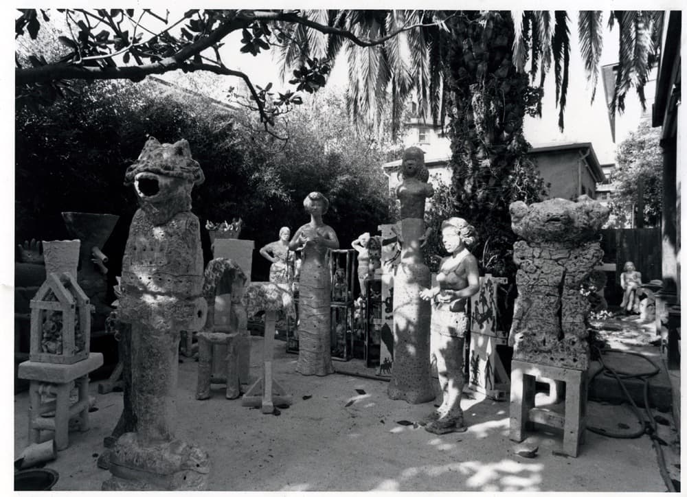 A concrete backyard filled with large ceramic sculptures of people, mythic creatures, and architectural stands, a large palm tree is in the center and shades the art.