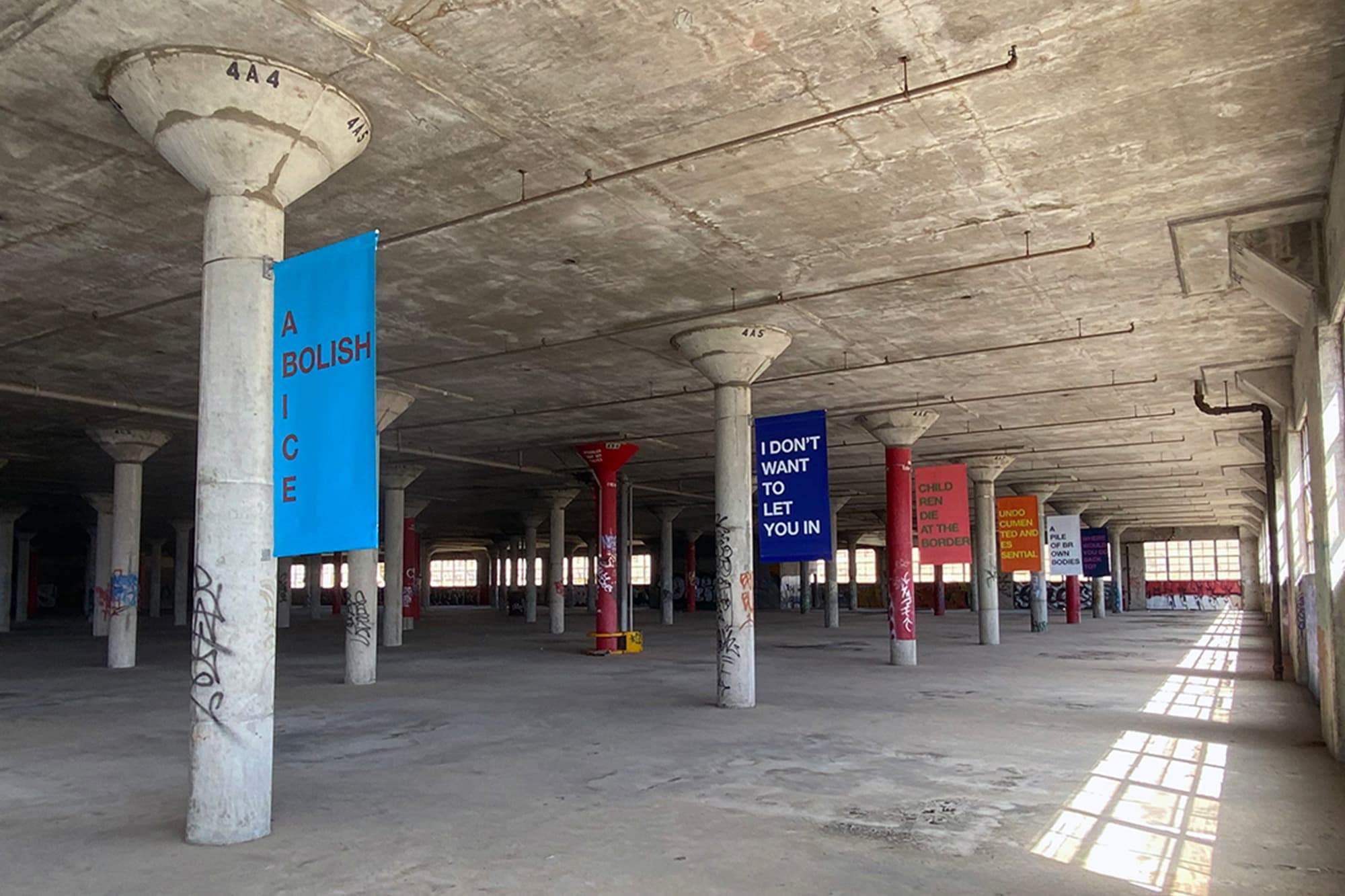 Warehouse like space with banners hanging from pillars. First banner reads: Abolish ICE.