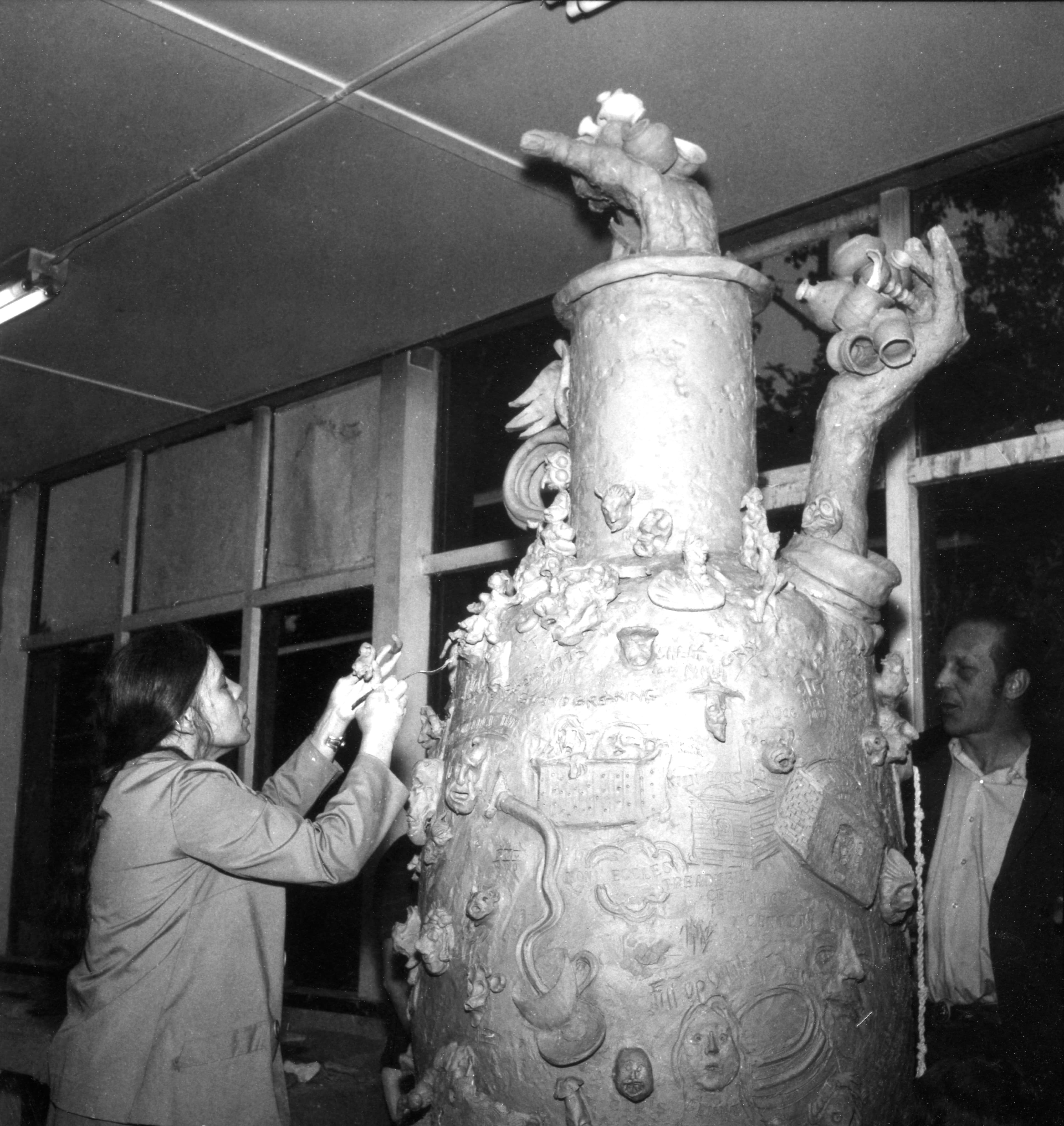 A woman adding a ceramic figure to a larger than life clay vessel decorated by two large sculptural hands and engravings.