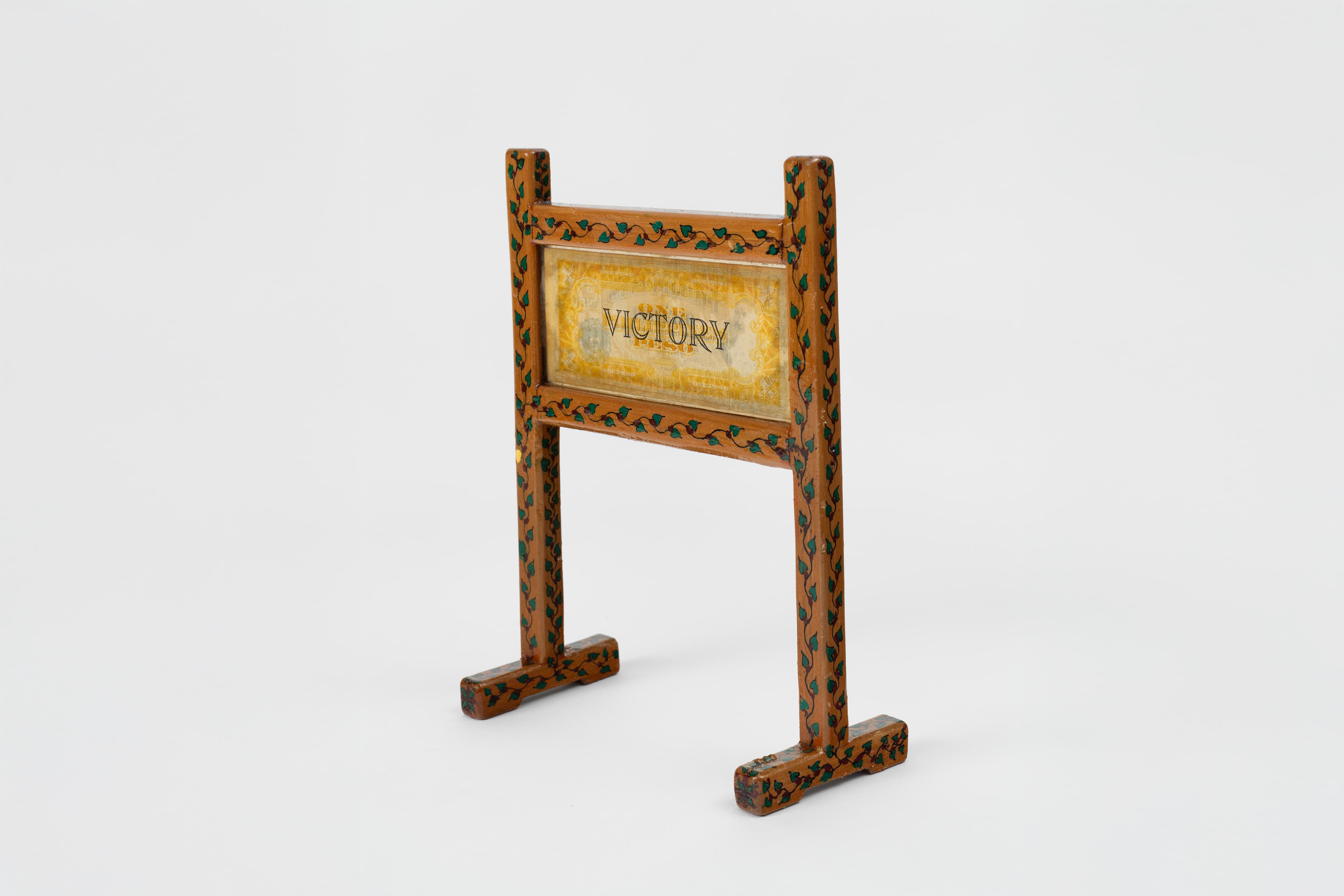 A wooden structure shaped like a freestanding billboard or post sign, angled slightly to the right against a plain white backdrop. The frame consists of two tall vertical posts connected by two horizontal crossbeams, all painted a warm tan color and decorated with a repeating pattern of hand-painted green ivy leaves and delicate vines. Suspended between the crossbeams is a rectangular, marbled yellow-and-white center panel with currency. Written in the currency is the word "VICTORY" in a clean, black, hand-drawn serif font.