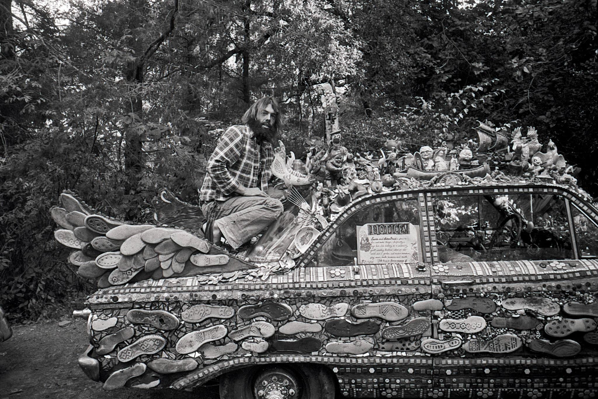 Man sitting on art car covered entirely in shoe soles, small figurines, and mosaic patterns. The man has long hair, a thick beard, and a plaid button-down shirt, he is perched atop the trunk of the car. The vehicle is parked outdoors next to a dense thicket of trees and woods, with a handwritten notice sign visible in one of the rear windows.