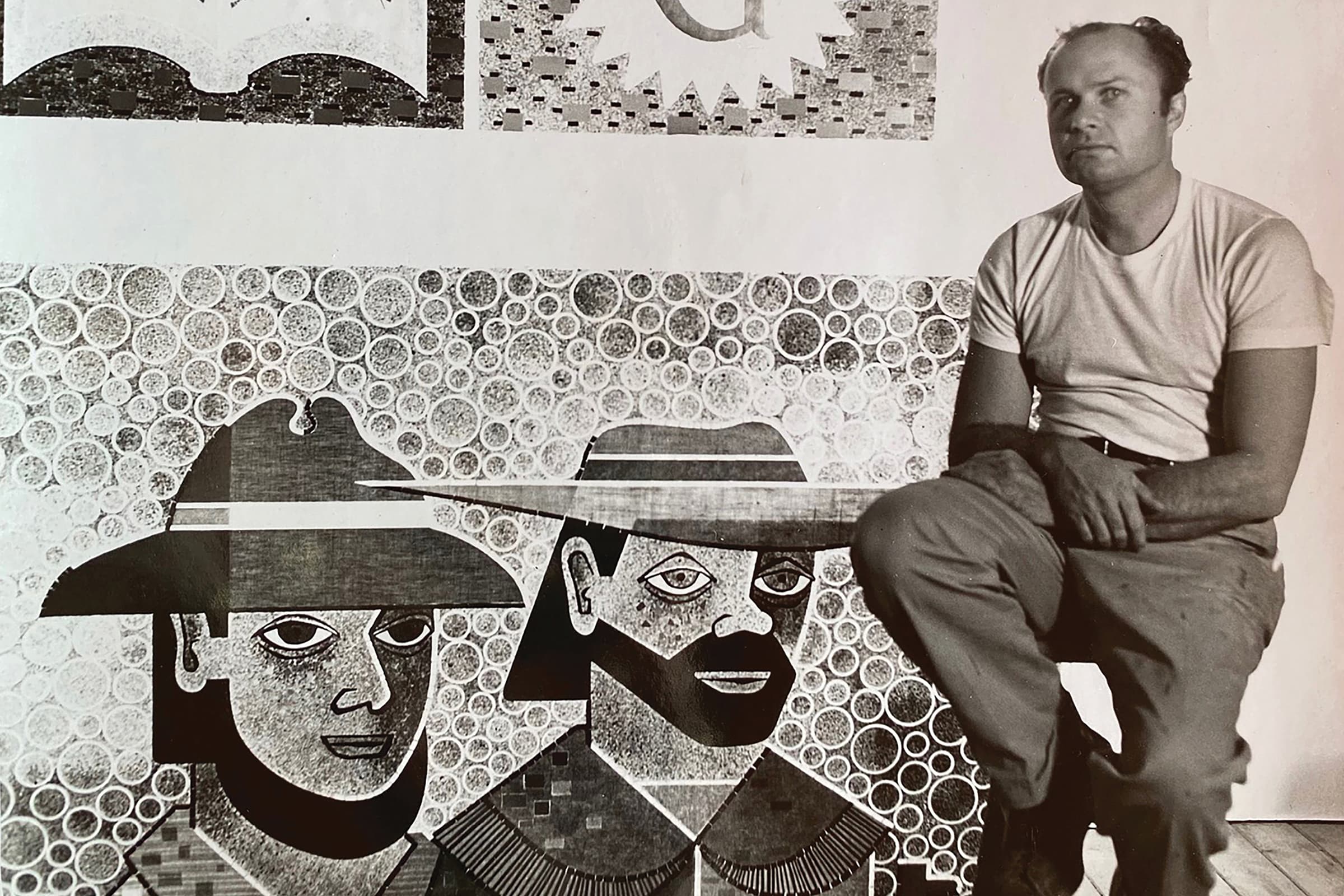 A black and white photograph captures artist Emile Norman posing beside a large, intricate multi-panel mosaic featuring portraits and abstract textures. Sitting to the right of the artwork on a stool, Norman is dressed casually in a light-colored T-shirt and dark trousers while looking directly towards the camera. The lower section of the mosaic next to him depicts two figures wearing wide-brimmed hats set against a background of densely patterned concentric circles, showcasing a complex blend of graphic detail and texturing.