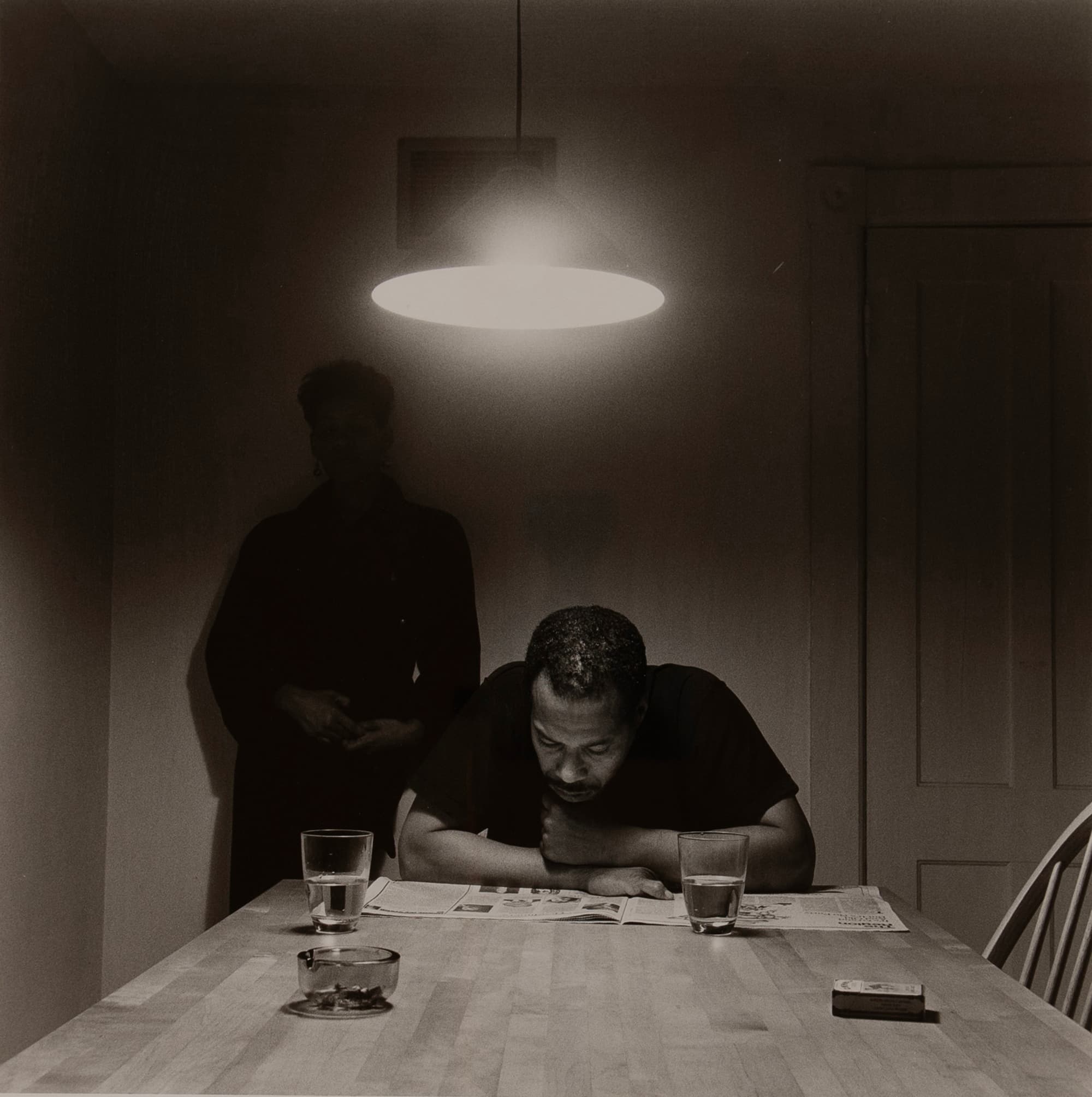 A black-and-white photograph of two people seated at a kitchen table beneath a hanging lamp. One person reads a newspaper while another stands behind them in shadow. Two glasses of water and an ashtray sit on the table.