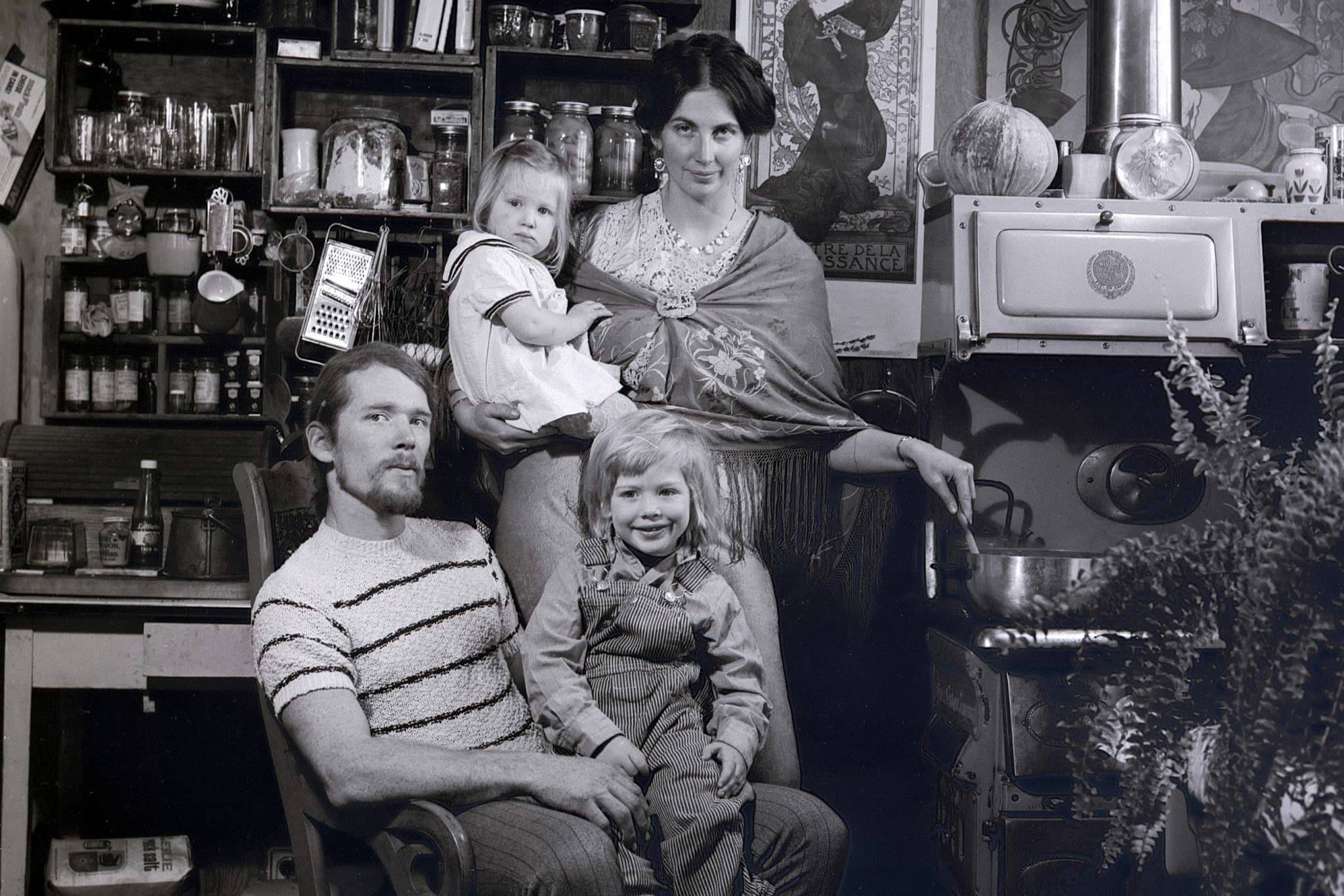 Family standing in kitchen. A woman stands by a vintage stove to the right, stirring a pot while holding a toddler in a light dress. In front of them, to the left, a man with a goatee sits in a wooden chair with a young child wearing denim overalls resting on his lap. The background features packed shelves of jars, glassware, and kitchen utensils.