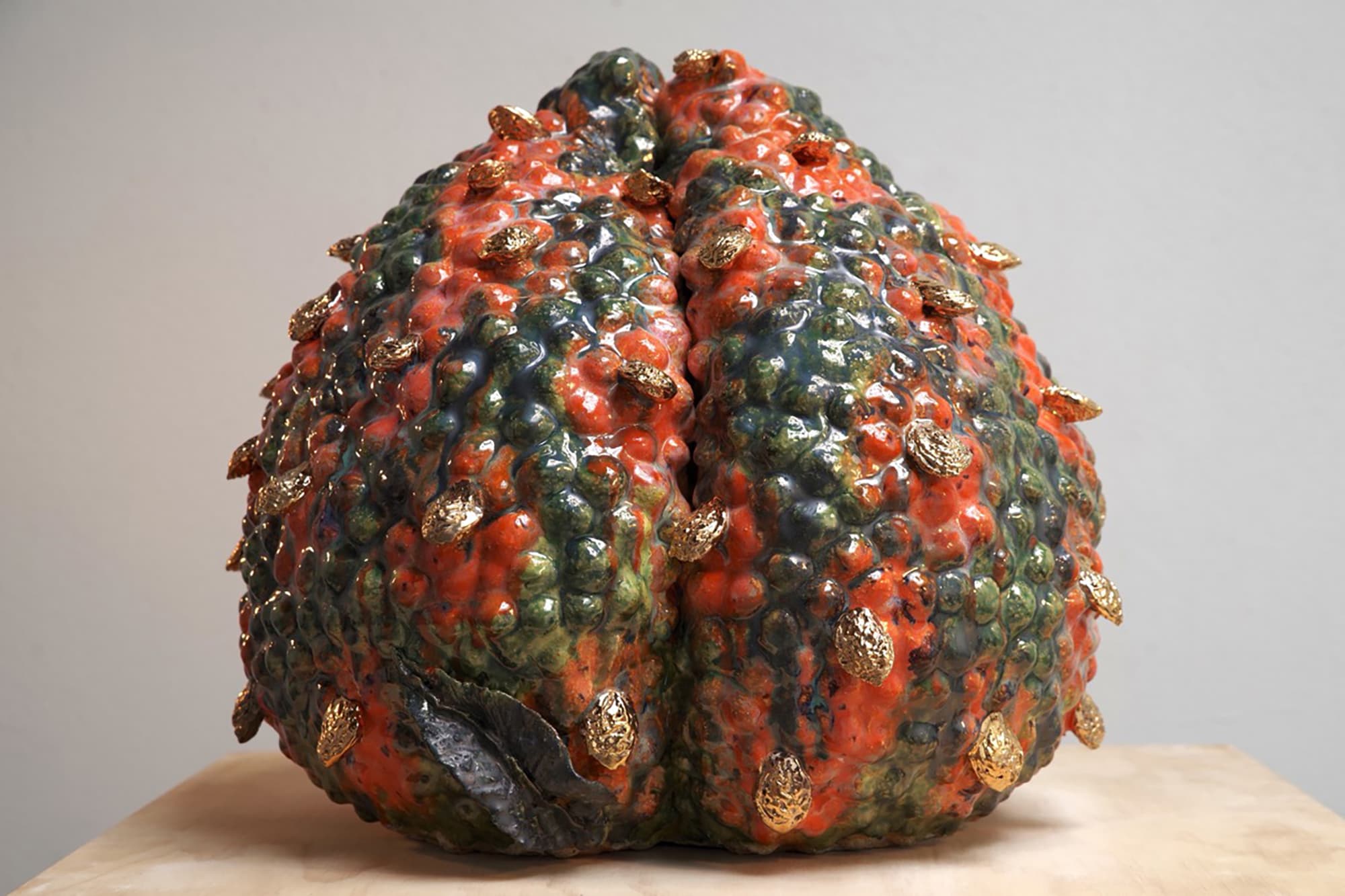 A textured, bumpy ceramic sculpture resembling a peach, resting on a light wooden surface. The piece features a high-gloss glaze blending shades of deep orange and mottled forest green across its uneven, dimpled surface. Dozens of small, textured metallic gold protrusions are scattered across the exterior, catching the light like small barnacles or gilded seeds. A single dark, metallic grey leaf-like shape sits tucked near the bottom left base of the sculpture.
