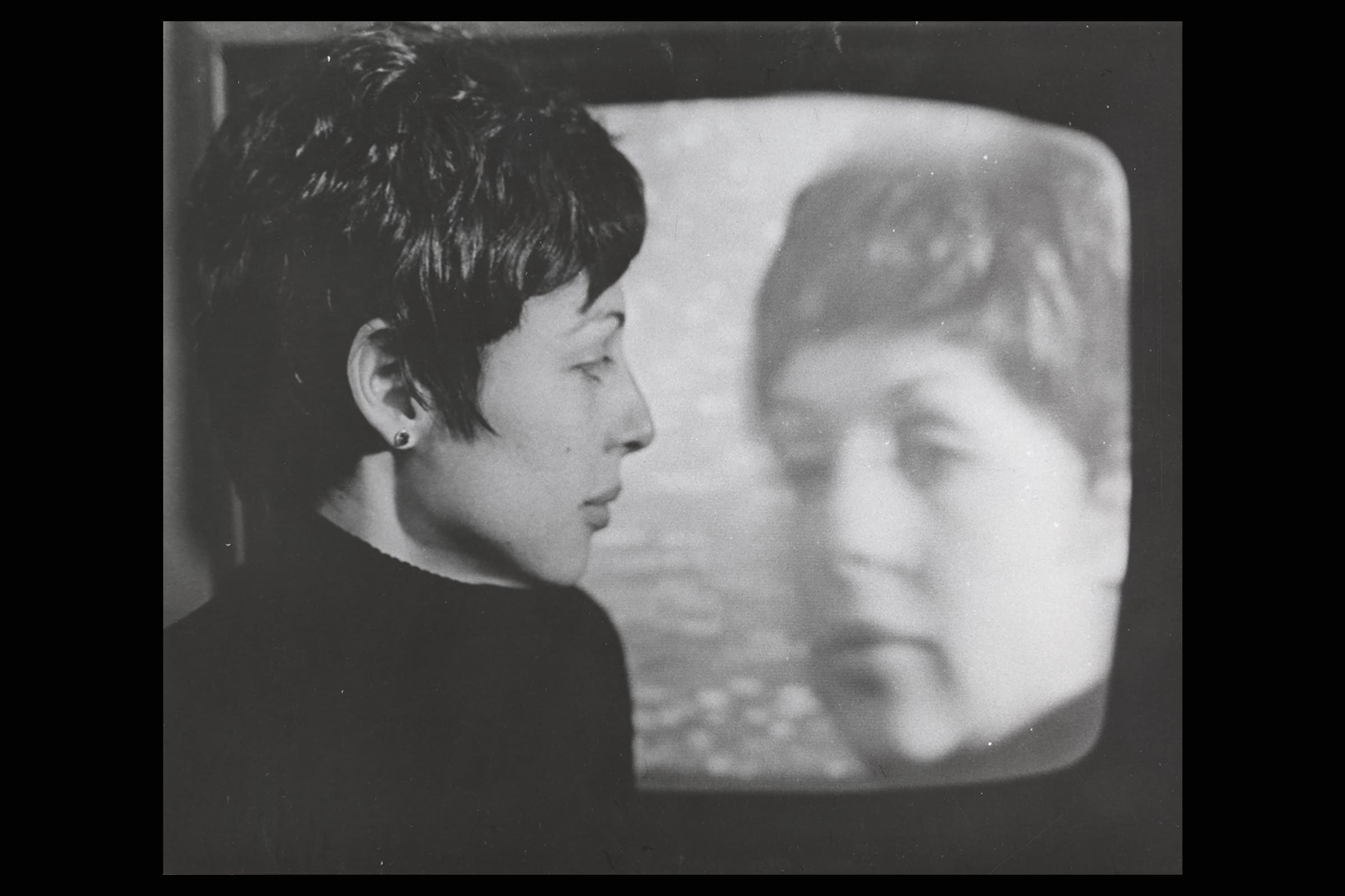 A person with short hair is looking at their image on a television screen. Both figures are in profile and the image is in black and white.