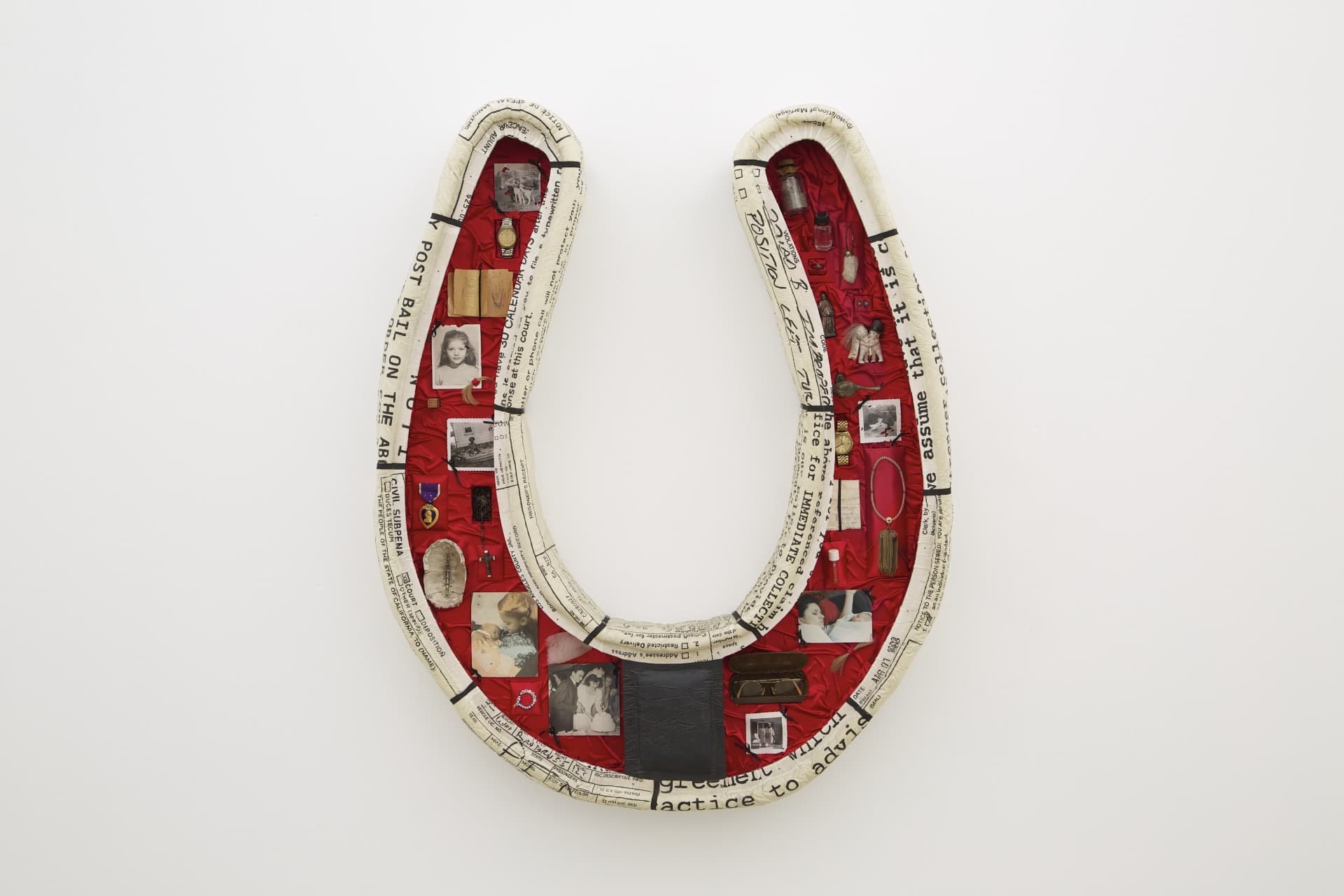 Horseshoe-shaped mixed-media artwork filled with photographs, keepsakes, and text fragments on a red fabric background.