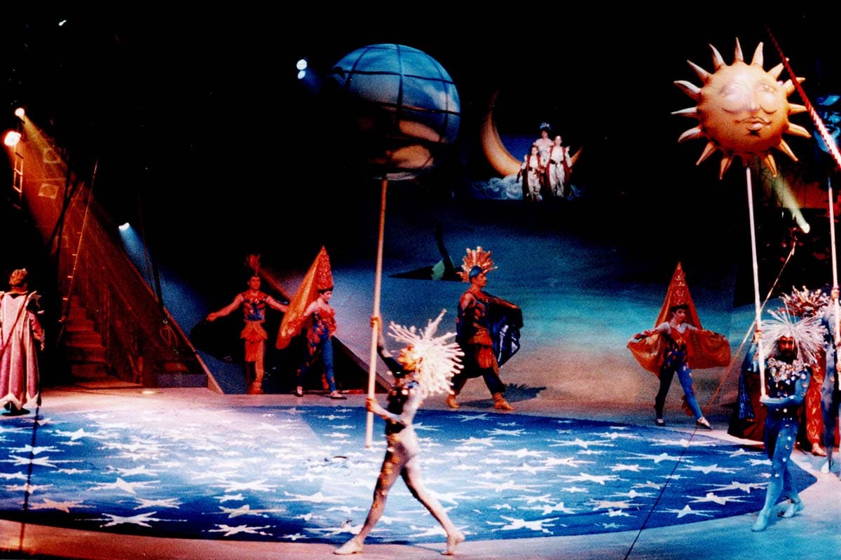 A wide stage production featuring actors in elaborate celestial-themed costumes. The stage floor is painted bright blue with numerous white stars. In the foreground, a performer wearing a white spiky wig and a patterned bodysuit walks while holding up a large globe prop on a long pole. To the right, another performer in a blue star-themed costume holds a pole with a smiling golden sun prop. Other characters in vibrant costumes stand or walk across the stage under a crescent moon structure in the background.