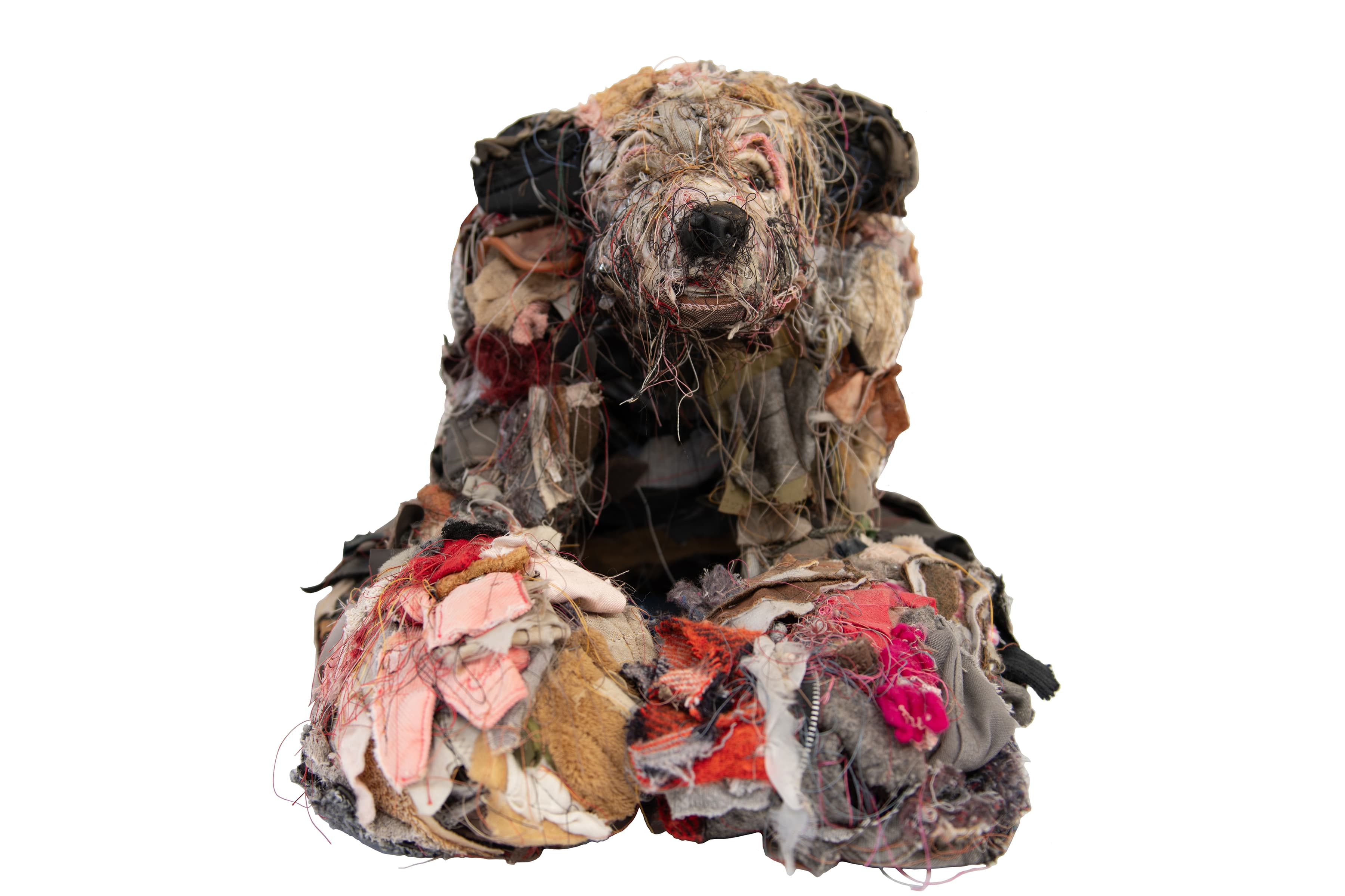 A sculpture of a seated dog made from layered pieces of cloth, fabric scraps, and loose threads. The dog faces forward with its head raised and its front paws resting on the ground.