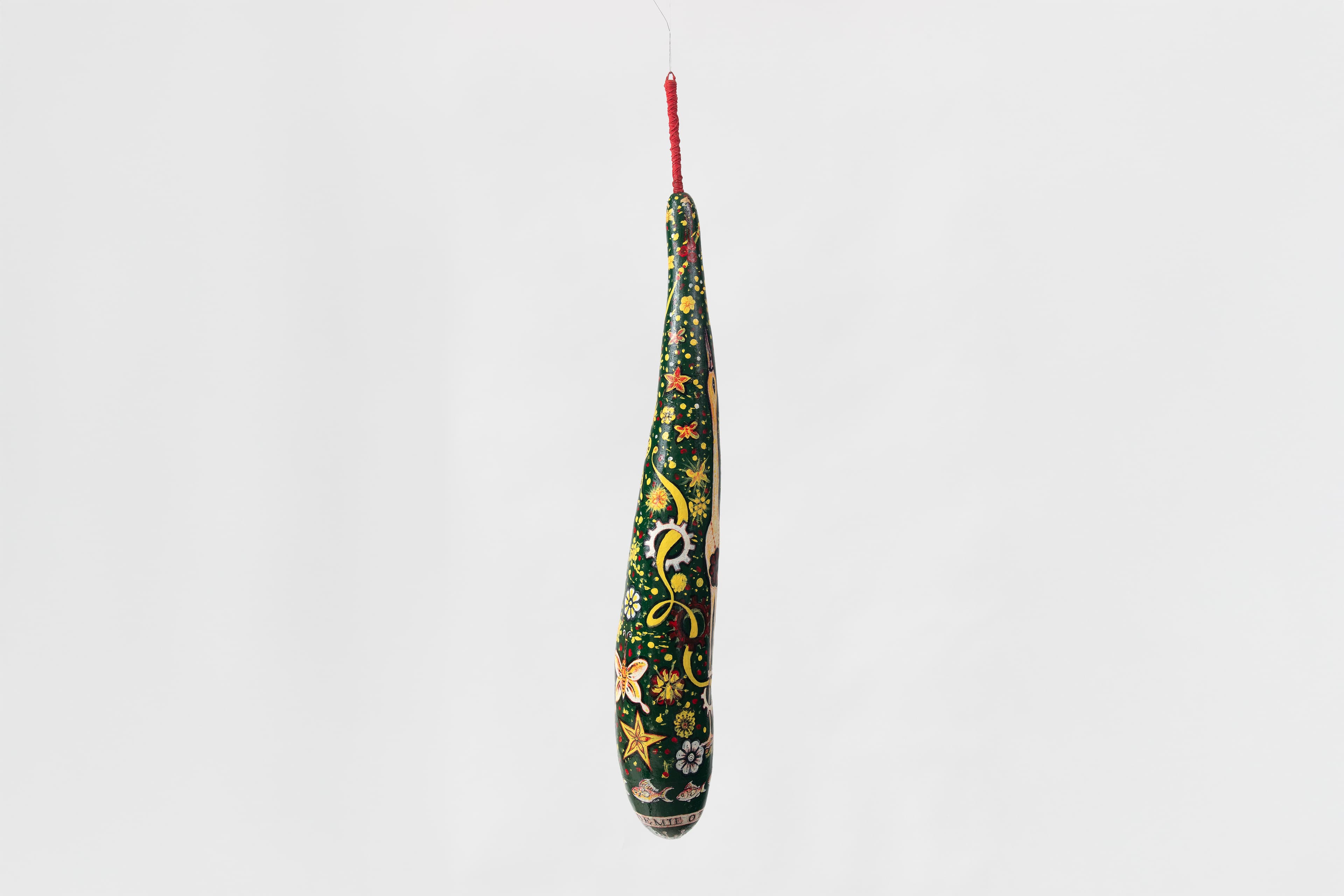 A single elongated, slender wooden gourd hanging vertically against a seamless white background, suspended by a bright red wrapped cord at its stem. The entire body of the gourd is painted dark forest green and covered in dense, highly detailed colorful illustrations. Swirling yellow ribbons, stars, and small multi-colored floral blossoms decorate the middle sections. Near the bottom, stylized orange-and-white fish swim above a painted banner framing the very base, which contains partially visible capitalized block text reading "EMIE OF".