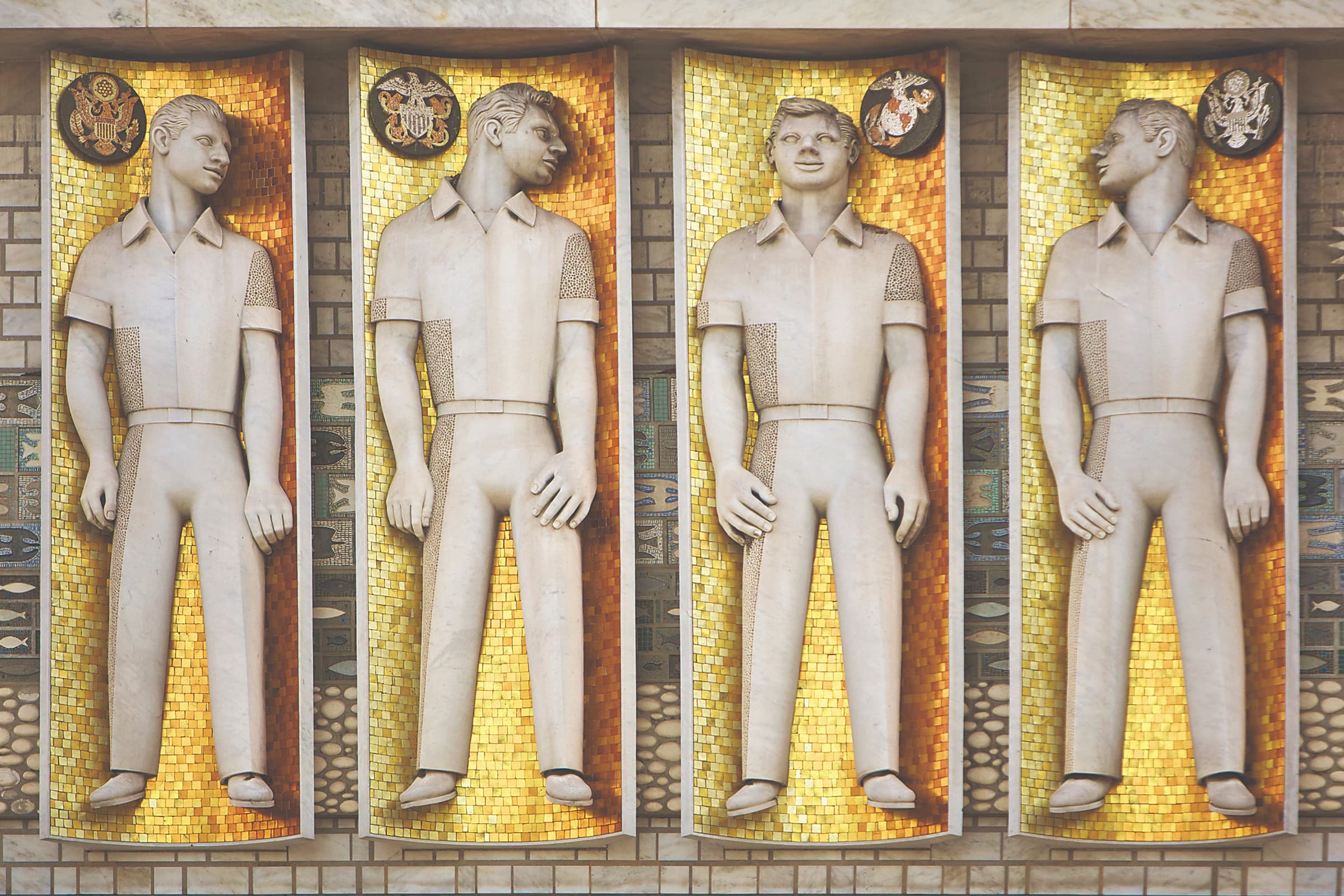 A bas-relief sculpture featuring four uniformed men standing inside gold mosaic alcoves. Each figure has a icon that refers to a branch of the United States Armed Forces, indicated by a circular military seal placed in the top corner of their respective alcove. From left to right, the branches shown are the Army, Navy, Marine Corps, and Air Force. The men are sculpted in a stylized, mid-century modern aesthetic, wearing collared short-sleeve shirts and trousers, with their heads turned at various angles. Geometric tiles and stylized symbols line the narrow pillars separating each section.