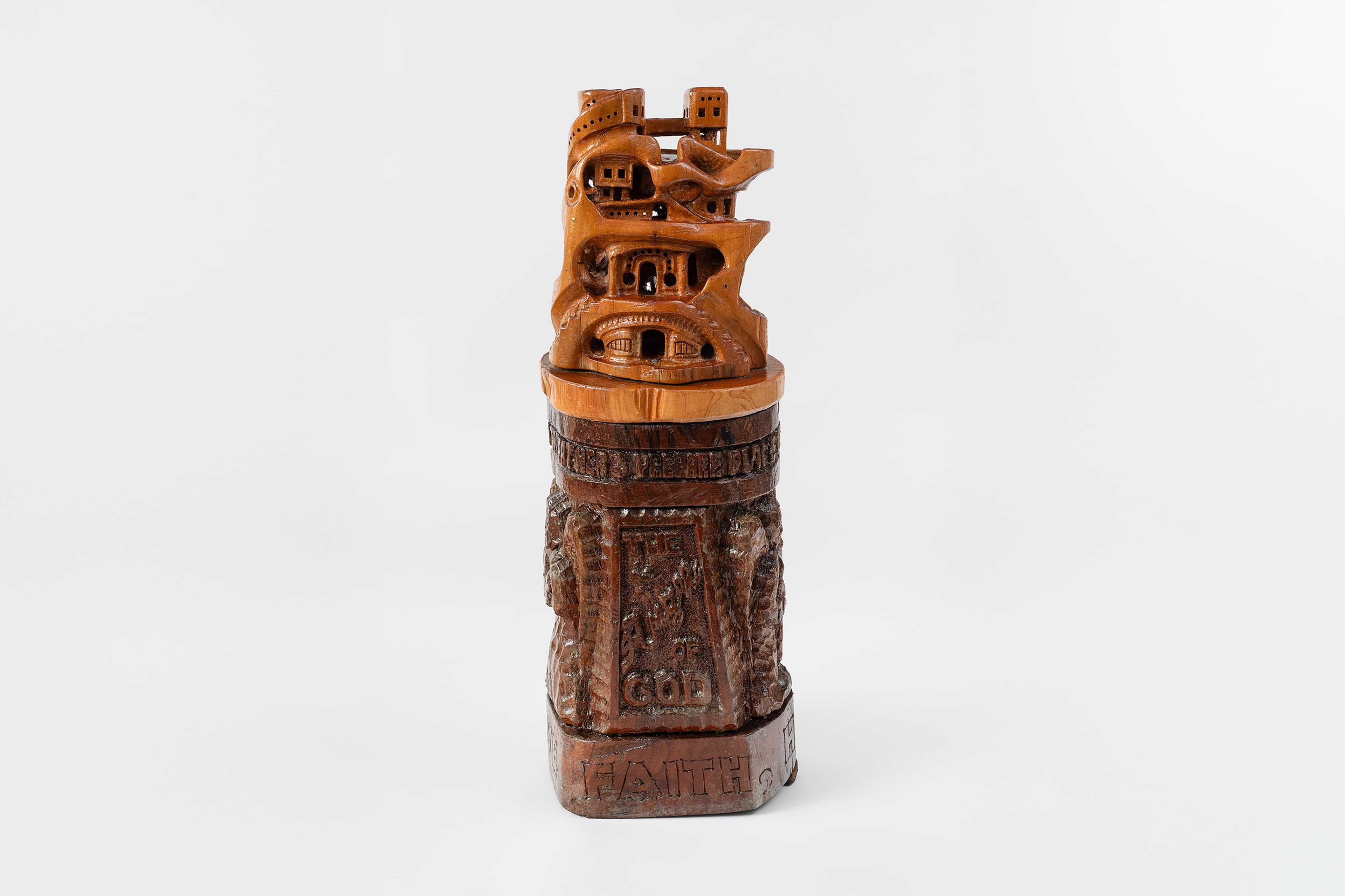 An intricate, multi-tiered cylindrical sculpture resting on a clean white surface. The piece is divided into distinct sections. The top third is carved from lighter, honey-colored wood, depicting a dense, miniature mountain citadel or architectural ruins with hollowed-out windows, archways, and bridges. This rests upon a larger, dark brown cylinder carved with dense, high-relief scenes of figures and landscapes. The front of this dark base features a prominent rectangular frame containing the carved text "THE HEAVEN OF GOD," which sits directly above the word "FAITH" carved in bold block letters across the bottom rim.