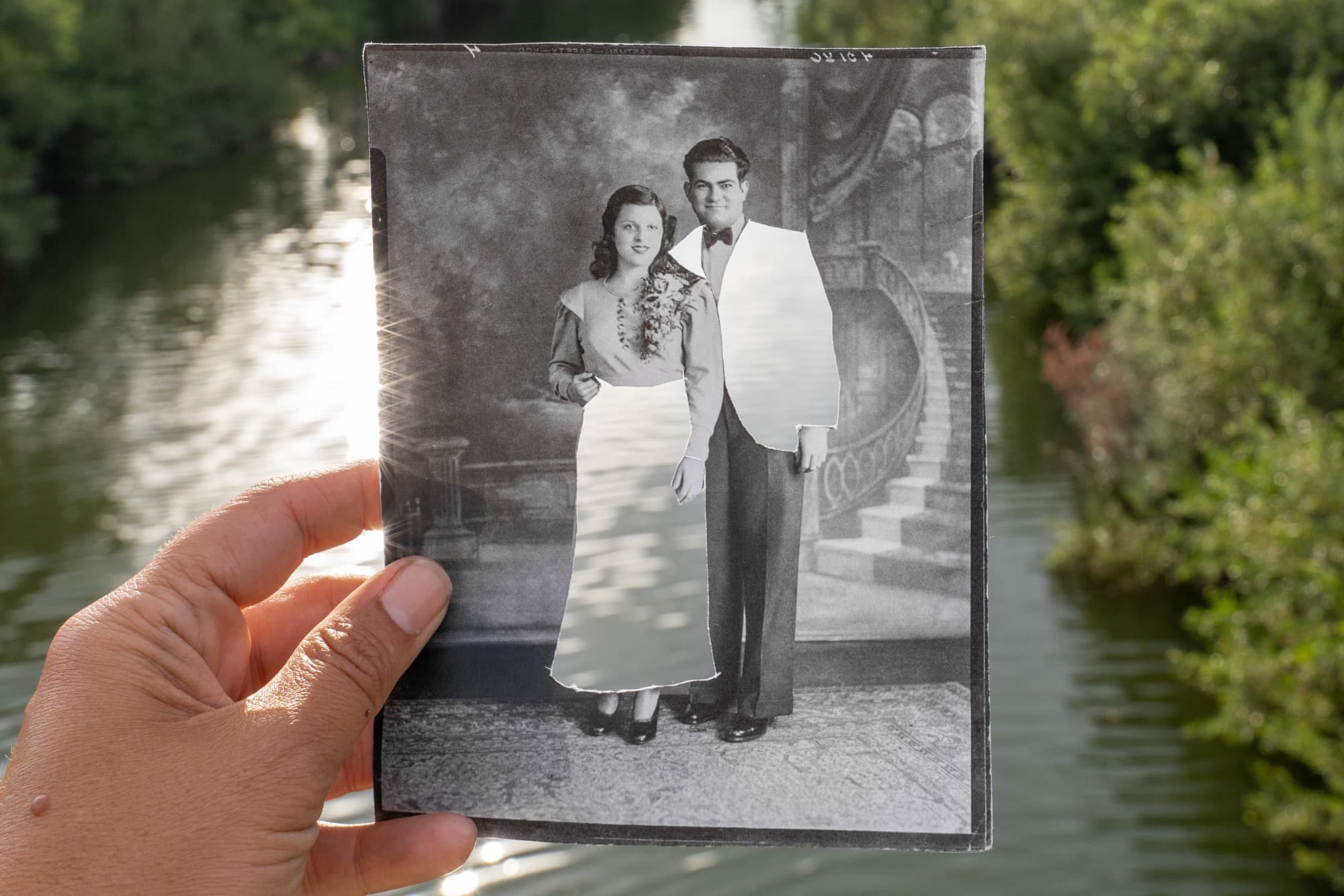 A hand entering from the lower left holds a vertical, black-and-white photograph in front of a blurred landscape of trees and water. The photograph shows a woman and man standing side by side before a painted backdrop. To the right, a carpeted staircase with a decoratively patterned bannister ascends to windows and drapery. A short stone barrier wall extends from the staircase and is partially obscured by the figures. The woman has long, dark hair and wears a blouse with real flowers cascading over one shoulder. The artist has cut away her skirt, leaving her feet in black high heels intact. The dark-haired man wears black dress shoes and slacks. His jacket has been cut away by the artist, leaving his white shirt and black bow tie intact. Through these openings, the landscape behind the photograph is revealed, including water reflecting sunlight.