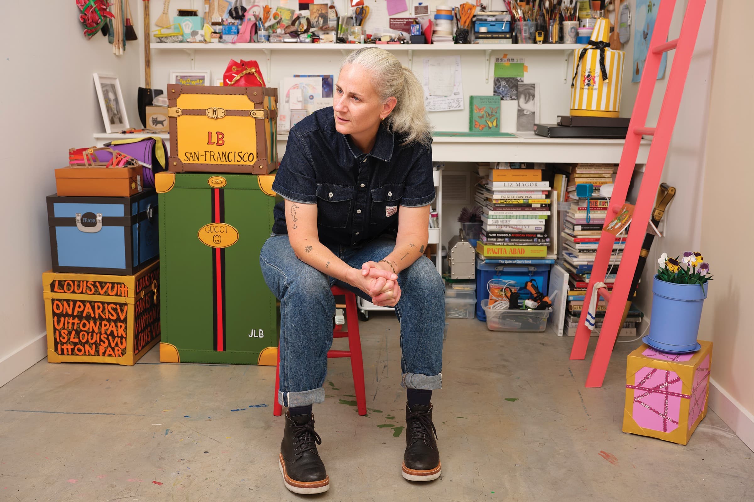 A person with light skin and grey hair tied back in a ponytail sits on a red stool in the center of a cluttered artist's studio. They are looking to their right, wearing a dark blue short-sleeved denim shirt, blue jeans rolled up at the cuffs, and black leather boots. Their hands are clasped together in their lap. In the background, a medium-size yellow trunk labeled LB SAN-FRANCISCO rests on top of a large green trunk labeled GUCCI. A stack of other stylized luggage sits nearby, including a blue trunk with a PRADA logo and a hand-painted LOUIS VUITTON box. To the right of the person, a tall stack of art books rests on the floor, including titles by Liz Magor and Pacita Abad. A bright pink ladder leans against the right side of the frame near a blue pot containing flowers. The background features white shelves packed with art supplies, storage containers, and various creative materials.