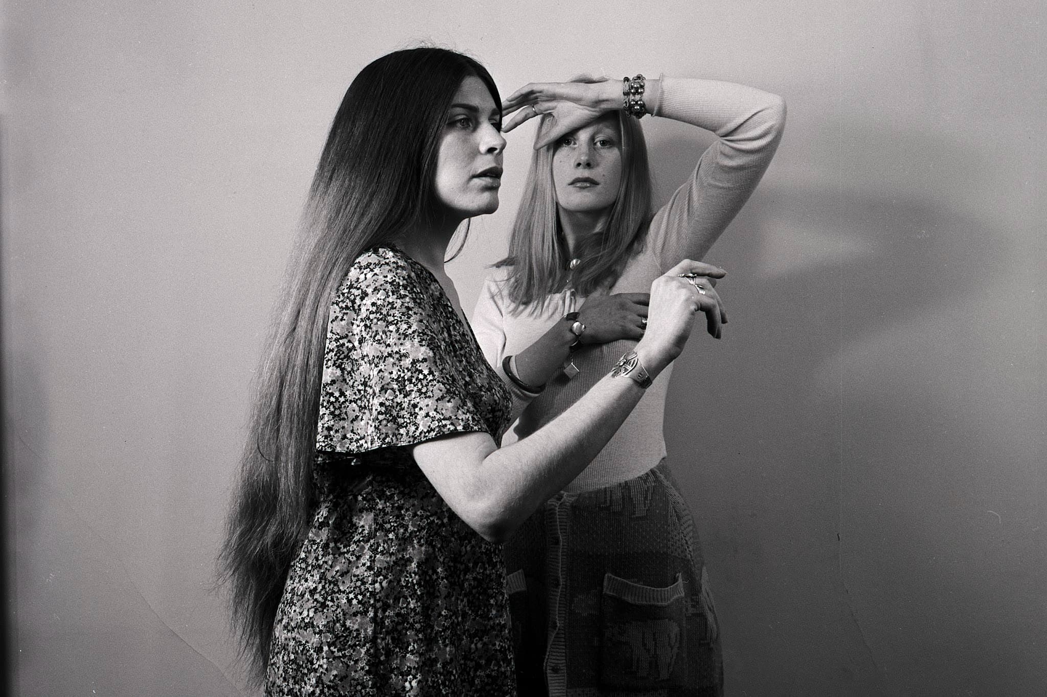 Two young women with long hair standing against a plain, light-colored wall. The woman in the foreground is in profile, sporting very long, dark hair and wearing a floral patterned dress. The woman behind her has blonde hair, wearing a light sweater and knit skirt, posing with one arm raised to shade her eyes and the other over her chest.