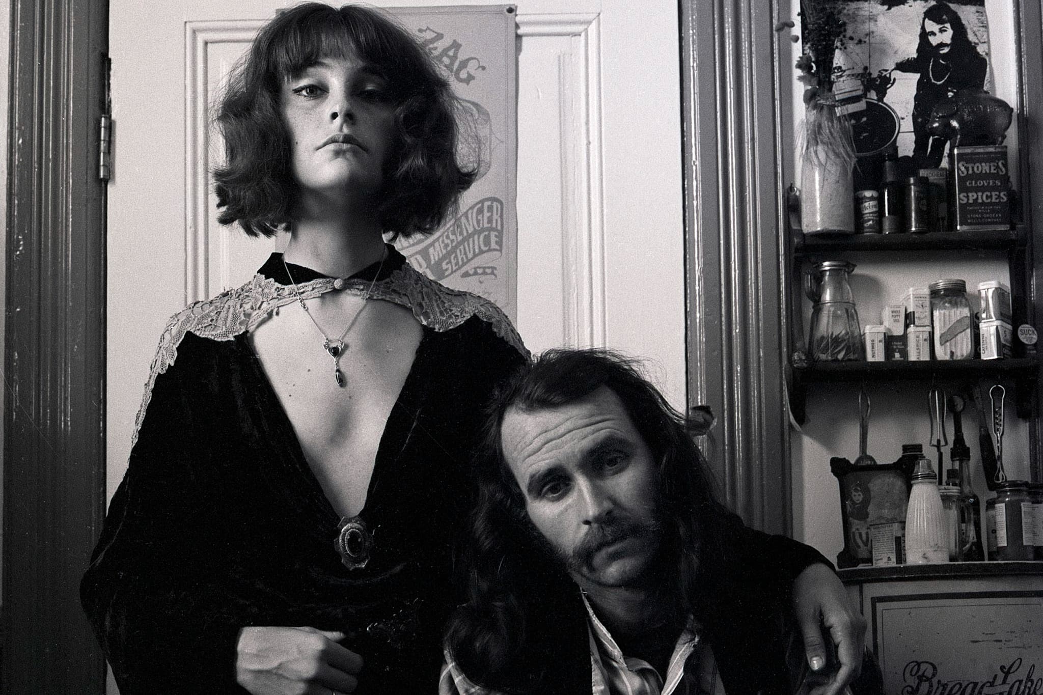Kathleen and Alton Kelley with child inside an apartment setting. Kathleen has a short, wavy bob haircut and she stands looking down towards the camera, wearing a dark velvet dress with a plunging neckline and lace trim. Alton wears long hair and a mustache and he leans his head against her hip, looking forward. In the background, posters are pinned to a door and a shelf filled with spice tins and bottles sits against the wall.