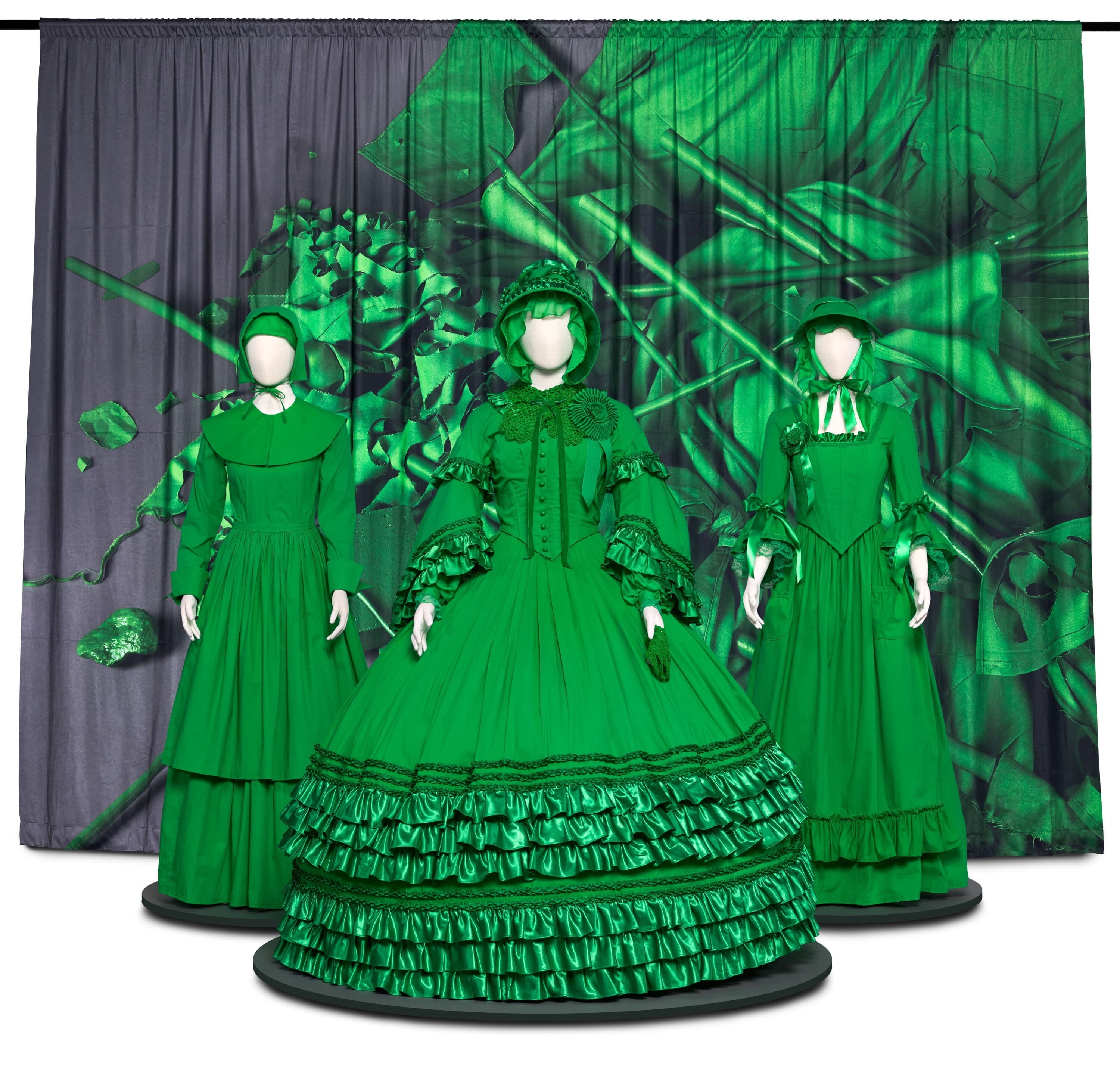 Three mannequins wearing bright green, historical-style dresses stand in a row in front of a large curtain backdrop. The central mannequin features a highly elaborate, wide-skirted Victorian-style gown with multiple tiers of ruffled fabric at the hem, voluminous long sleeves, and a matching bonnet. Flanking it on either side are two mannequins wearing simpler, long-sleeved green gowns with bonnets. The background consists of a heavy curtain printed with a distorted, abstract pattern of bright green shapes and lines layered over a dark grey base.