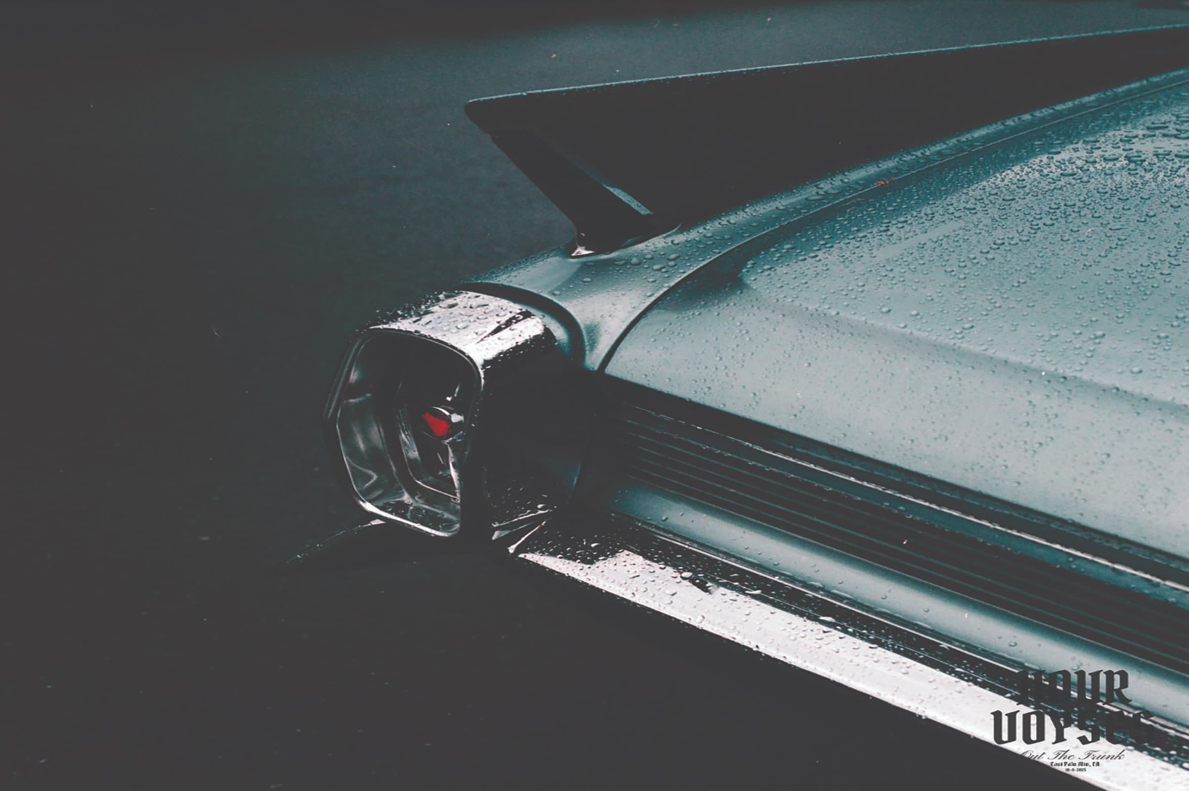A photograph showing the rear of an aqua blue car, covered with drops of rain.