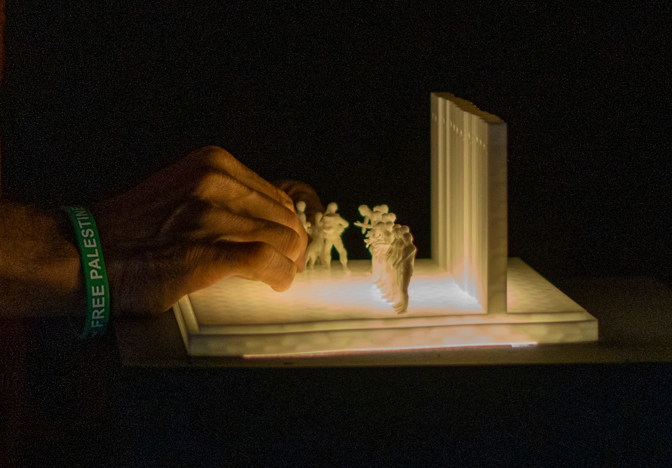 A close-up shot under low light captures a hand wearing a green wristband with the text "FREE PALESTINE" adjusting small, white, sculpted figurines on an illuminated white rectangular base. The figurines represent a line of people standing opposite a large vertical white wall structure, with warm light glowing upwards from the base.