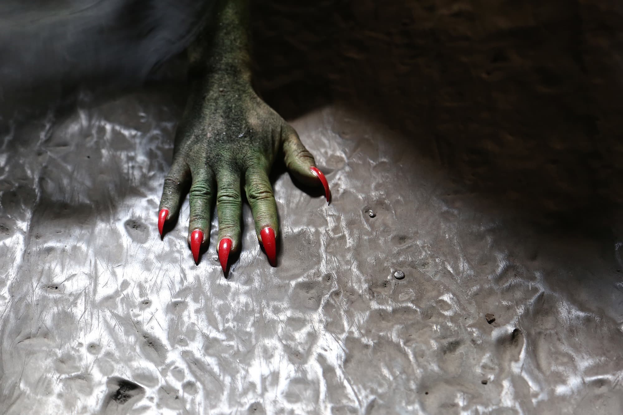 A green hand with long red nails on textured stone.