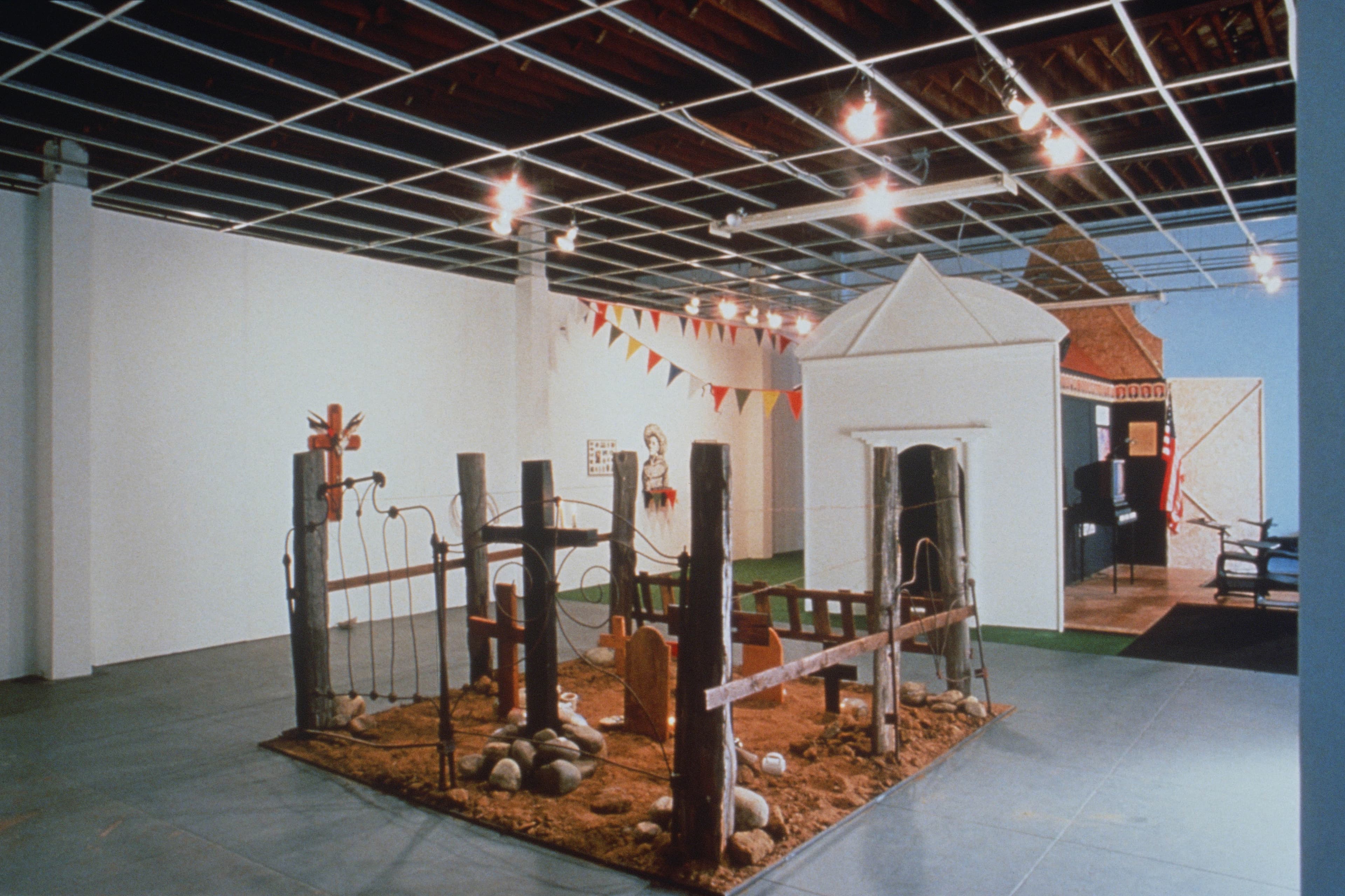 An installation view composed of a rectangular dirt plot on the floor containing wooden posts, crosses, a rusted metal gate, and barbed wire, resembling a graveyard or fenced enclosure. In the background, a white architectural structure resembling a small white mausoleum or house stands next to a walkway leading toward a darker section with an American flag. A string of multicolored triangular pennant flags hangs high across the white gallery walls connecting to the house.