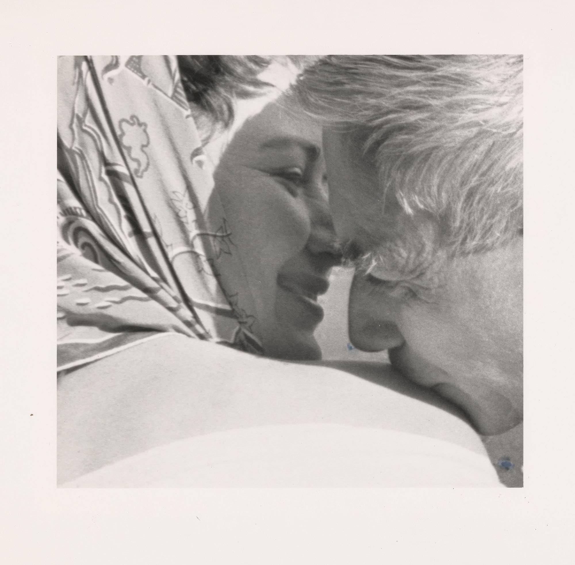 Photo framing the faces of two people and shoulders, to the left a woman turns her head to the right while the face of a man is leaning towards her right shoulder, he kisses it.