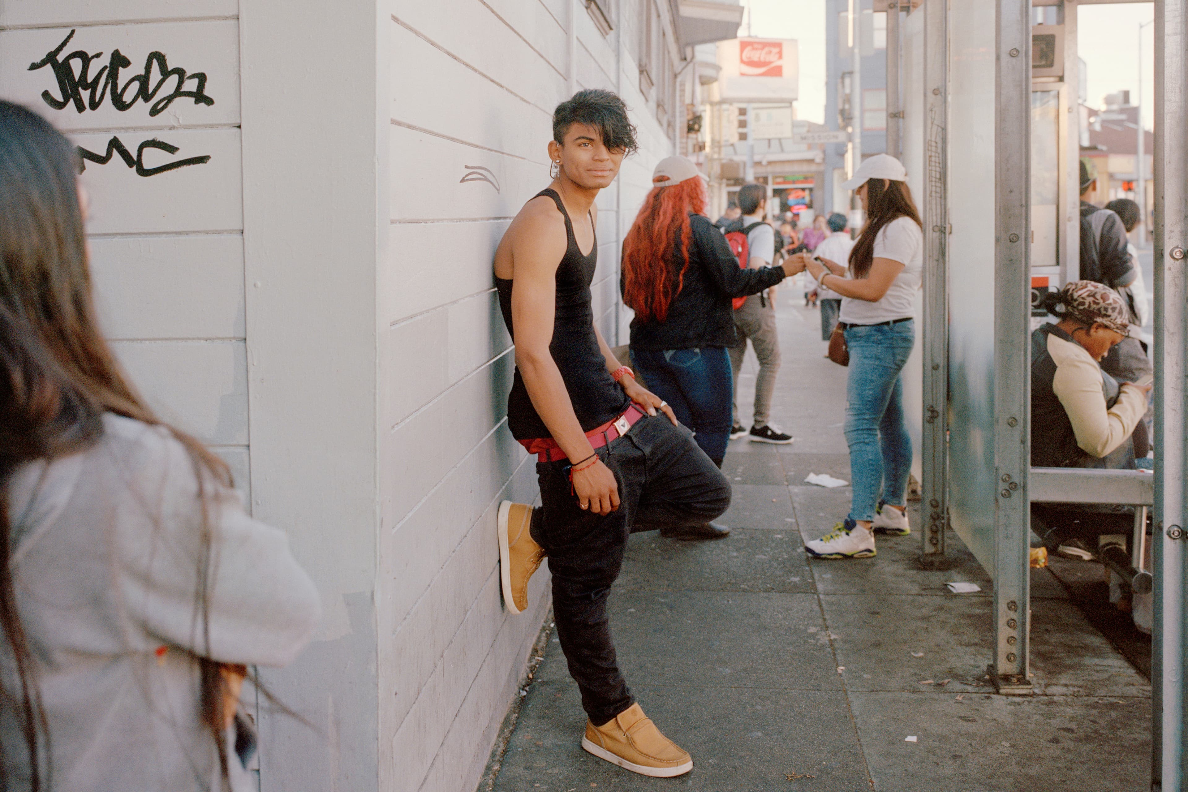 Boy with dark hair and dark clothing leans against a white wall in San Francisco's Excelsior District. In the background are other people waiting for the bus. He is the only person looking directly at the camera.
