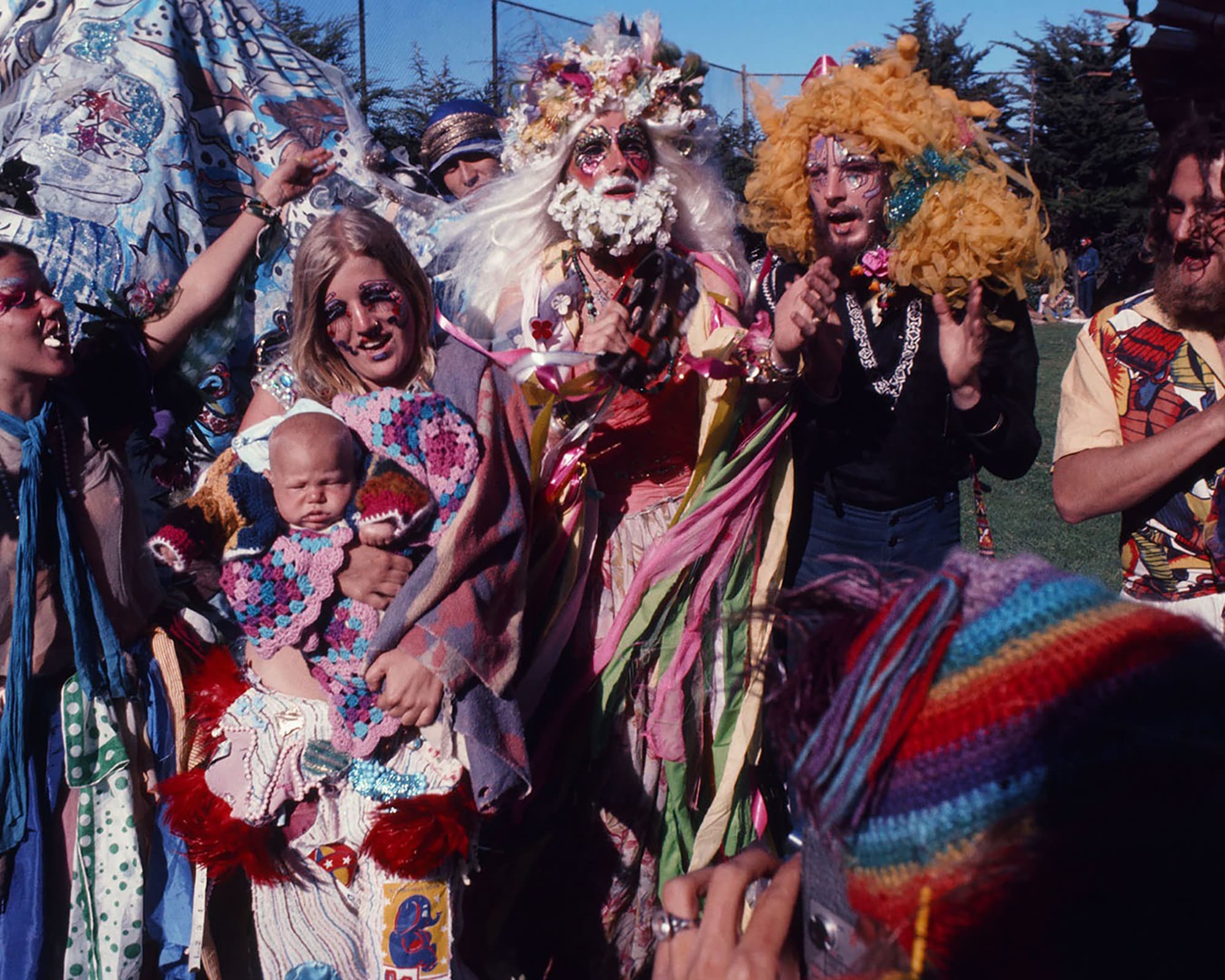 Photograph of members of The Cockettes theater group in eccentric, colorful costumes celebrating outdoors in a park. Near the center, a person wears an elaborate white beard and crown made of what could be flowers, surrounded by long, flowing pastel ribbons. To their right, a a blonde person in heavily patterned makeup holds a baby wrapped in a pink and purple crocheted blanket. To their left, a person with bright blue face paint wears a large, voluminous yellow yarn wig and a black shirt decorated with pearls. Other individuals in eclectic, bohemian clothing and patchwork fabrics stand around them on a green lawn, while a person in a rainbow-striped knit hat captures the scene in the foreground.